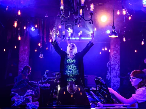 Performer on stage with outstretched arms, surrounded by musicians and illuminated by colorful stage lights and hanging lamps.