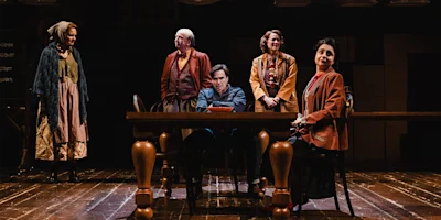 Five actors in period costumes perform on a stage with a wooden table and chairs, under theatrical lighting, suggesting a dramatic scene in a play.