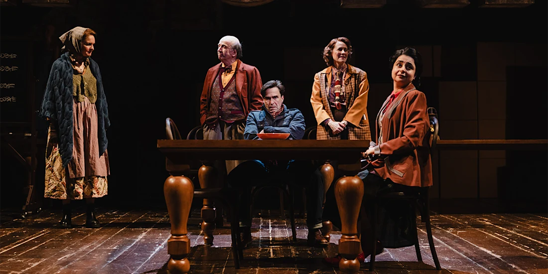 Five actors in period costumes perform on a stage with a wooden table and chairs, under theatrical lighting, suggesting a dramatic scene in a play.