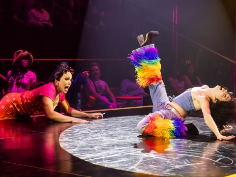 Two performers are on stage. One is on the floor with their legs extended upwards wearing rainbow-colored leg warmers, while the other is watching with a surprised expression. People watch from the background.
