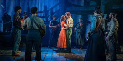 A man and woman embrace on a wooden stage, surrounded by musicians in period-style clothing under blue stage lighting.