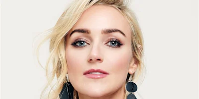 A woman with light skin, blue eyes, and blonde hair looks at the camera. She is wearing large, dark statement earrings and has shimmery eye makeup and pink lipstick.