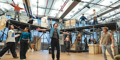 A group of performers rehearses on a multi-level stage set indoors, with one person standing center front with arms outstretched, while others are spread across platforms and stairs.