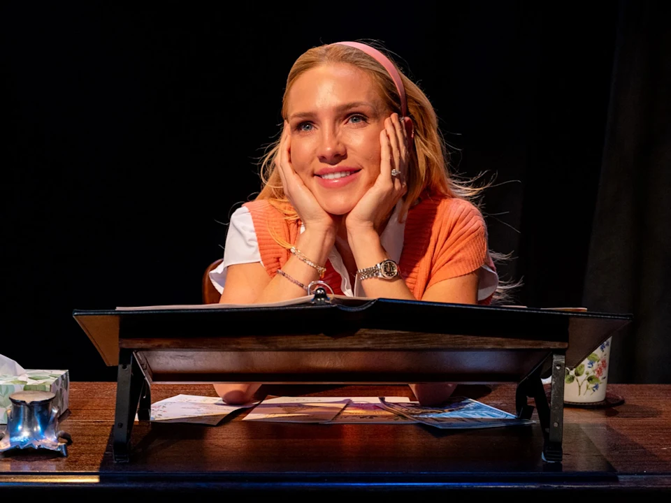 A woman with blonde hair and a pink headband sits at a wooden desk, smiling with her hands on her cheeks, looking upward. Papers, a tissue box, and a mug are on the desk.