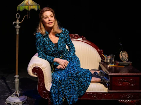 A woman in a blue patterned dress sits on a vintage white and wood sofa next to a lamp and a side table with a framed photo, glass, and books, against a dark background.