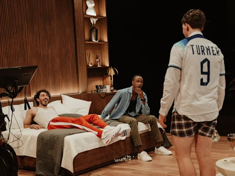 Three men are in a modern bedroom; one sits on the bed, one sits at the foot, and one stands wearing a soccer jersey and boxers with “Turner 9” on the back.