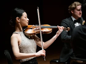 Production photo of The Chamber Music Society of Lincoln Center: Summer Evenings VI, showing a man in formal attire plays the cello while a woman in a silver dress plays the violin in a concert setting.