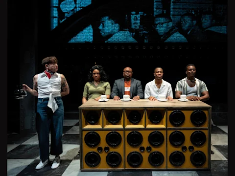Five people sit at a counter with a speaker front, each with a cup, while another person stands holding a coffeepot; black-and-white historical photos are projected on the wall behind them.