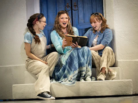 Three women sit on steps, smiling and looking at a book together. They are dressed casually, with a blue door in the background.