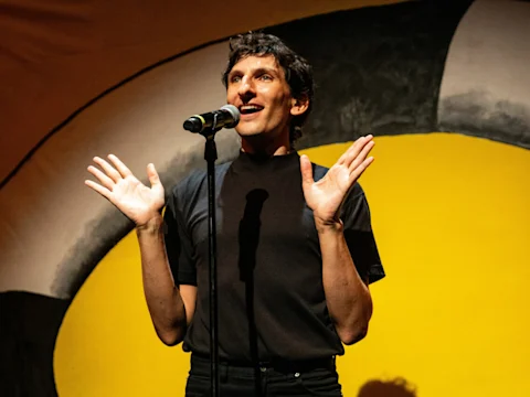 Photo of Morgan Bassich stands onstage in front of a microphone in Can I Be Frank?