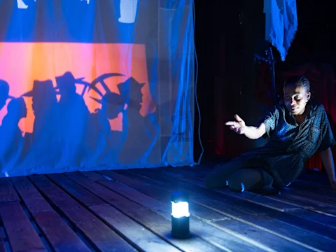 A woman sits on a wooden floor next to a lantern, gesturing toward a lit screen displaying silhouettes of people holding tools.