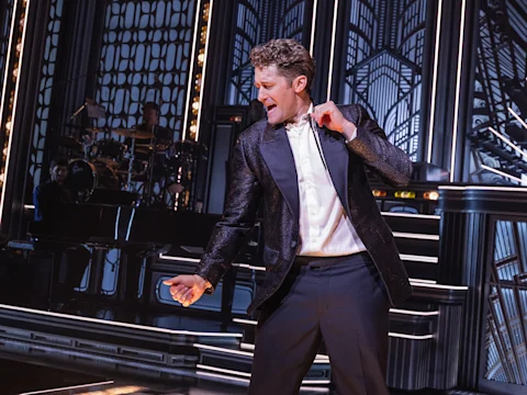A man in a dark tuxedo jacket and white dress shirt performs on stage in front of a live band.