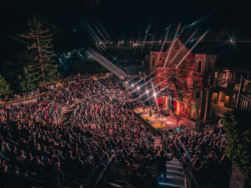 A large audience watches a nighttime outdoor performance at an amphitheater. Stage lights illuminate the performers, and beams of light extend across the image. Trees and a building are visible in the background.