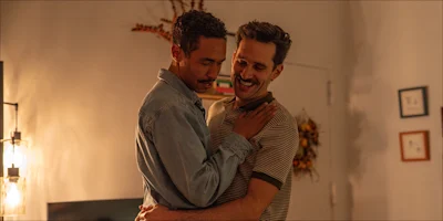 Two men embracing and smiling at each other in a dimly lit room.