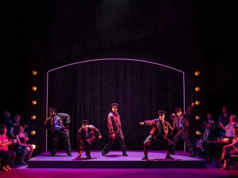 A group of performers in coordinated outfits are dancing energetically on a stage with purple lighting, surrounded by an audience seated on both sides.