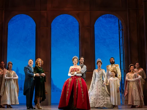 A group of actors in elaborate period costumes stand on stage in front of large arched windows with a snowy backdrop, performing a theater production.