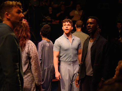 A group of people stand closely together in a dimly lit room, with one man in a light blue shirt and trousers looking forward while others face different directions.