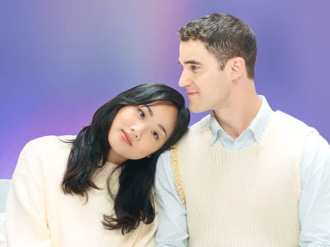 A woman with long dark hair rests her head on the shoulder of a man with short brown hair. Both are wearing light-colored sweaters and sitting against a purple background.