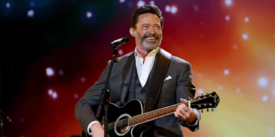 A man in a suit plays a black acoustic guitar and sings into a microphone on stage with colorful lights in the background.