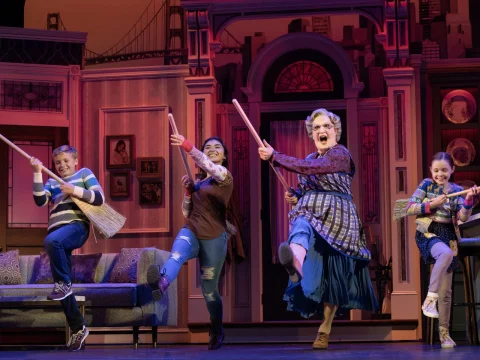 Four performers in colorful costumes energetically dance with brooms on a stage set resembling a living room, featuring a couch and backdrop of a cityscape.