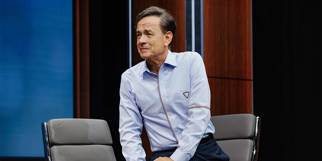 A man in a blue dress shirt sits on the edge of a gray chair in a modern, wood-paneled room.