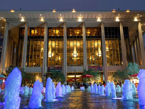 Production photo of Renee Fleming In Recital in Los Angeles showing,Illuminated fountain with blue lights at the foreground, with a modern building displaying large glass windows and bright interior lights in the background.