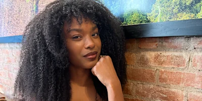 A woman with curly hair leans her chin on her hand, sitting indoors against a brick wall and a large framed photograph.