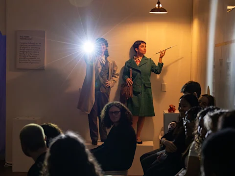 Two women dressed in vintage clothing stand on a platform, one holding a flashlight, in a dimly lit room with a seated audience in the foreground.