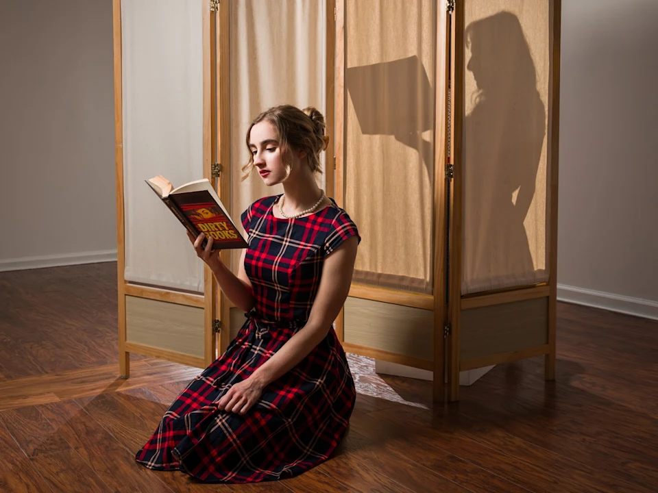 A woman in a plaid dress kneels on a wooden floor reading a book, with her shadow visible on a folding screen behind her, also holding an open book.