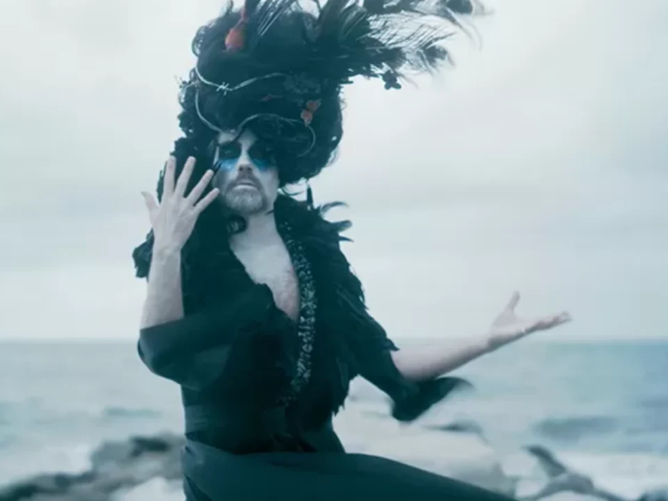 Person wearing an elaborate black feathered headdress and costume poses dramatically near a rocky shoreline.