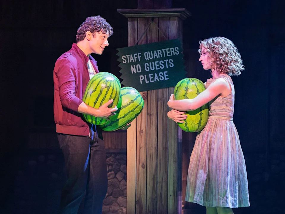A man and a woman in 1960s-style clothing hold watermelons while standing on either side of a wooden post with a sign that reads “Staff Quarters No Guests Please.”
