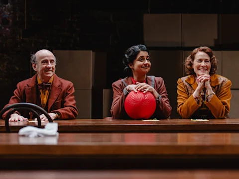 Three adults sit at a wooden table, two women and one man, all smiling. The woman in the center holds a red rubber ball. Cardboard boxes are stacked in the background.