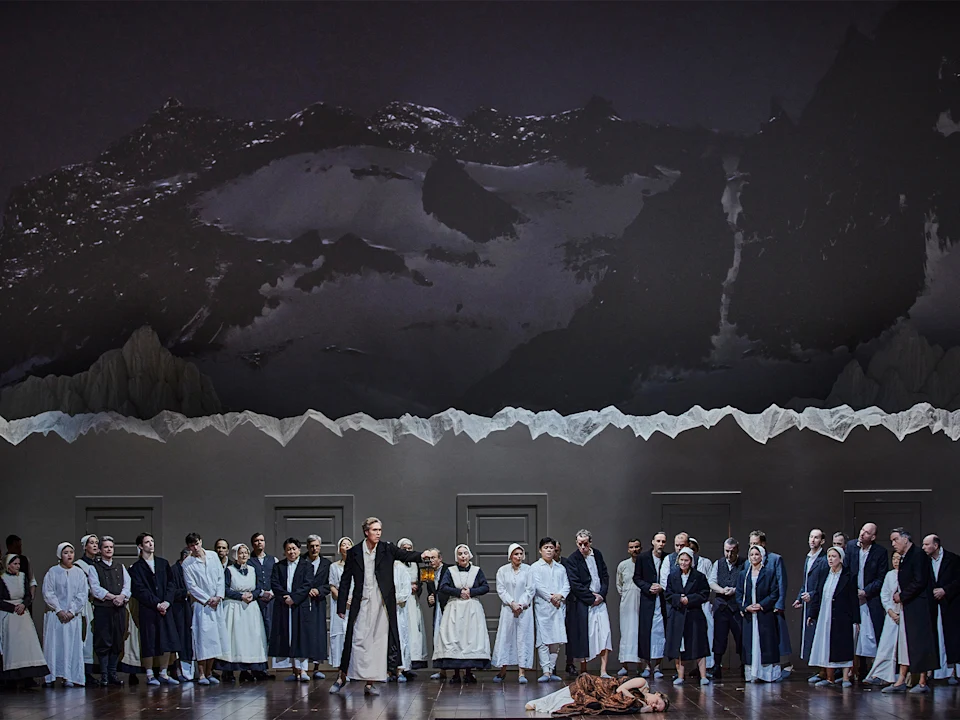 A large group of people in white and dark robes stand on a stage with mountain scenery in the background; one person lies on the floor in front.