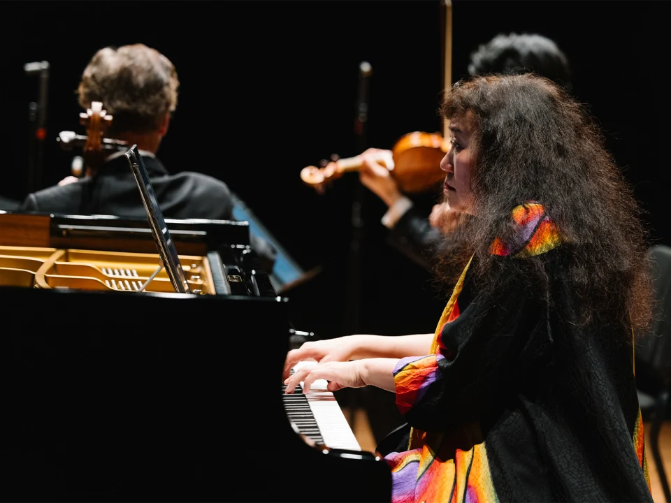 A woman plays the piano on stage, with a violinist and other musicians in the background. She is wearing a colorful dress.