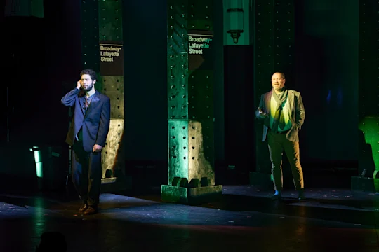 Two men in suits stand on a dimly lit stage, each near a pillar labeled "Broadway-Lafayette Street," with one man holding a phone to his ear.