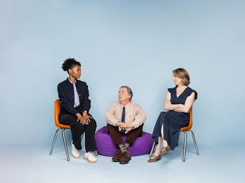 Three adults are seated against a light blue background; two women sit on orange chairs while a man sits on a purple bean bag between them.