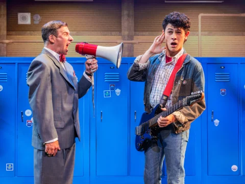 A man in a suit shouts through a megaphone while a young man holding a guitar stands nearby, covering one ear. Blue lockers are in the background.