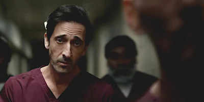 A man in burgundy scrubs stands indoors, looking directly at the camera, with two other people blurred in the background.