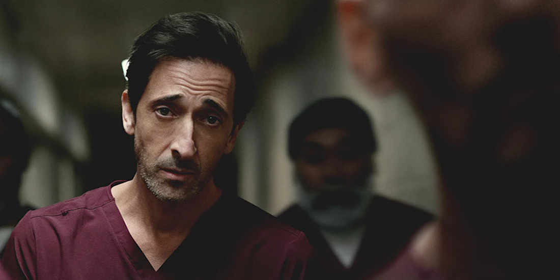 A man in burgundy scrubs stands indoors, looking directly at the camera, with two other people blurred in the background.