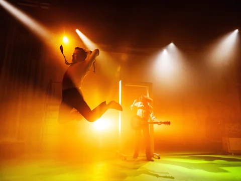 A person jumps in mid-air with arms raised while another person plays guitar on stage, both illuminated by dramatic orange stage lights.