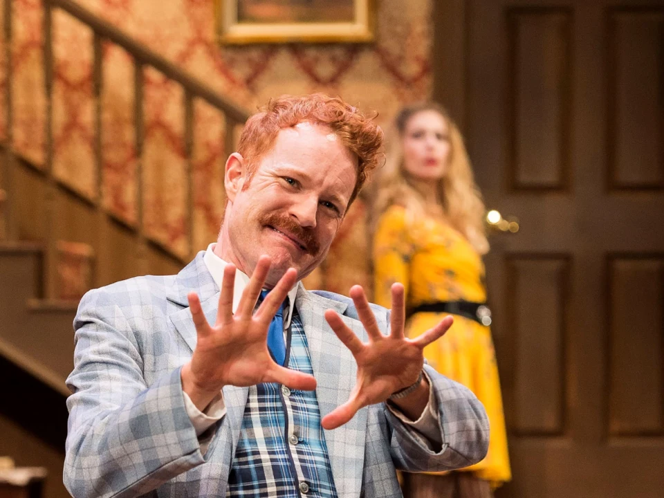 A man with red hair and a mustache in a plaid blue suit gestures animatedly with his hands while a woman in a yellow dress stands in the background by a staircase.