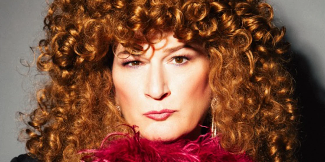 Person with thick, curly red hair and bold makeup, wearing large earrings and holding a pink feather boa, looking directly at the camera with a neutral expression.