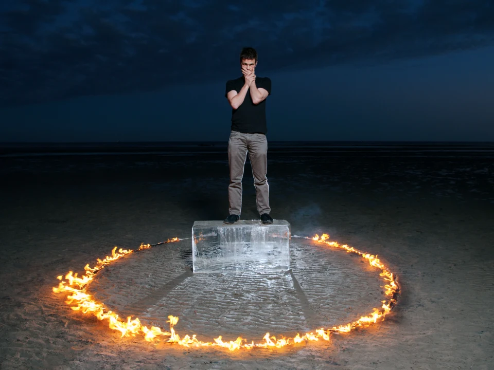 Production photo of Deep History in New York City, showing a man stands on a large block of ice surrounded by a ring of fire on a flat, barren landscape at twilight.