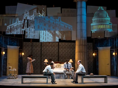 Four people in mid-20th century attire are on a stage set resembling a hotel room, with projected images of buildings and the Capitol dome above them.