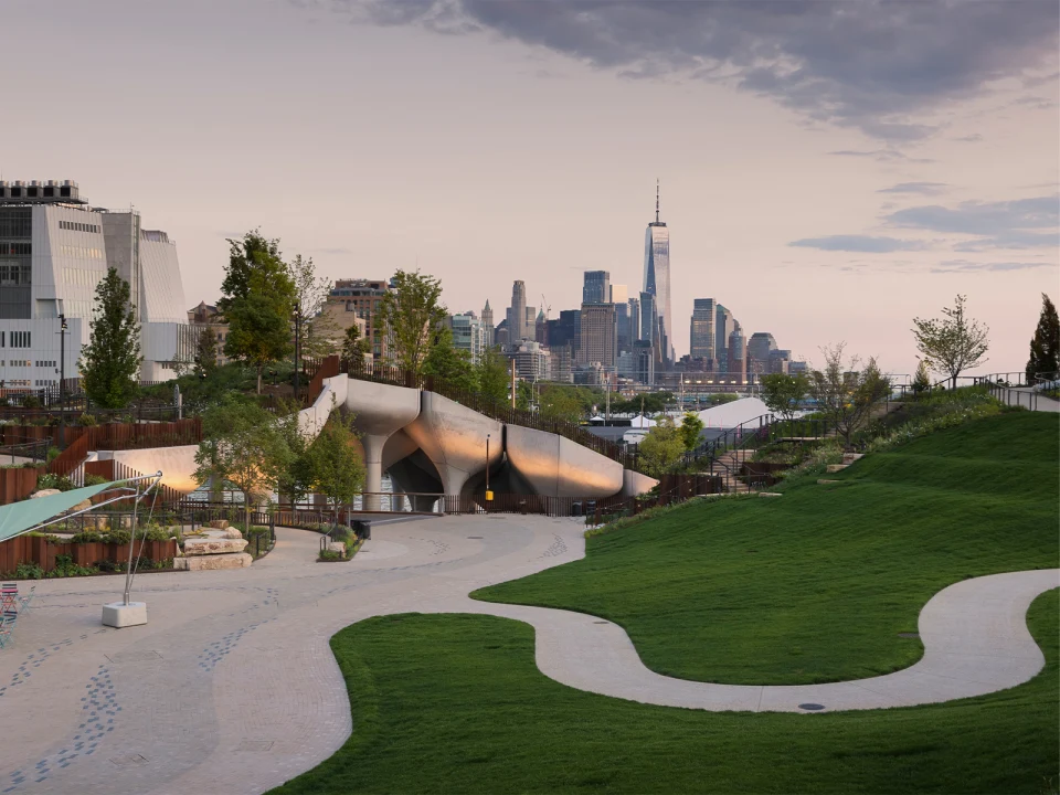 View of a modern park with a curvy pathway and green grass leading to an innovative pavilion, with a city skyline featuring skyscrapers in the background at dusk.