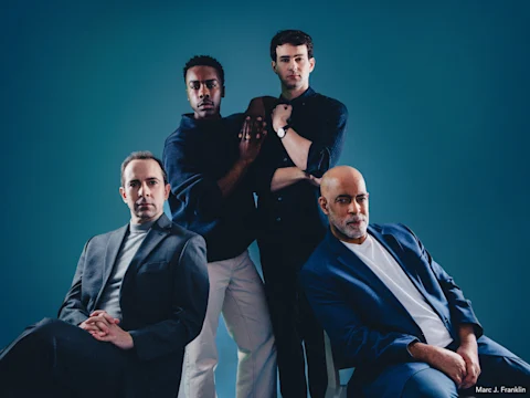 Four men pose against a blue background; two are seated in front wearing suits, and two stand behind them, one in a dark shirt, the other with arms crossed.
