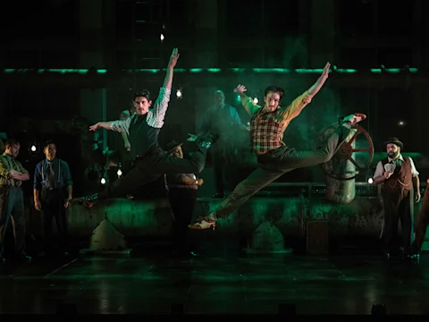 Two men leap in mid-air with arms and legs extended on a dimly lit stage, while several others watch in the background.