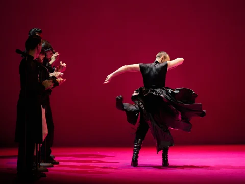 A dancer performs energetically in the center of a dimly lit stage with a vibrant pink background, while three people observe and clap from the side.