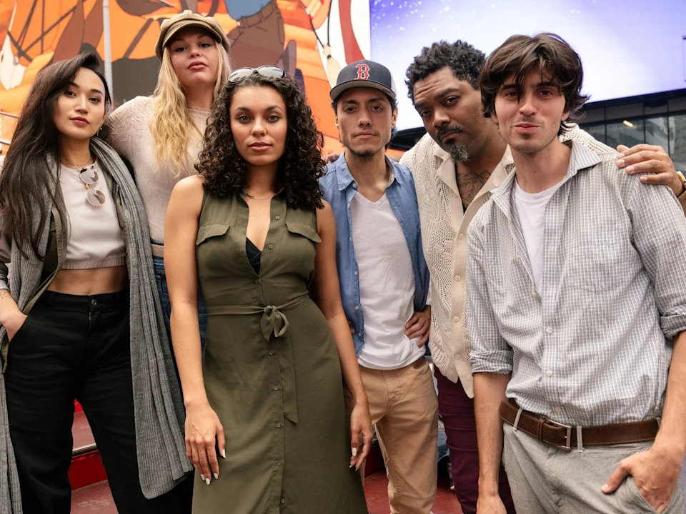 Six people pose together outdoors in casual clothing, standing closely in front of a colorful mural.