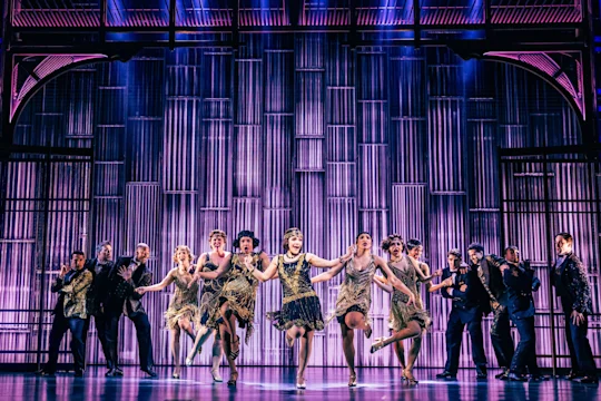 A group of performers in 1920s-style costumes dances energetically on stage, with dramatic purple lighting and a metallic backdrop.